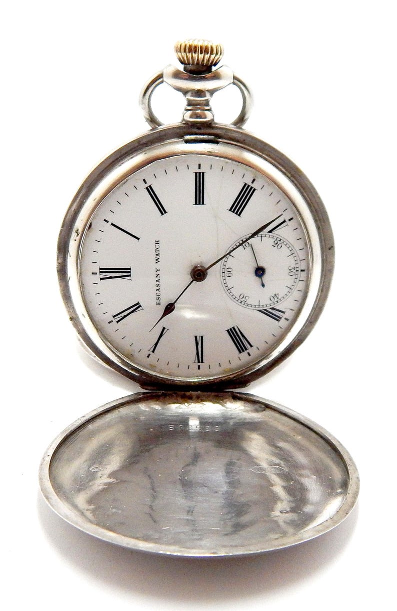 May include: A silver pocket watch with a white face and black Roman numerals. The watch has a small seconds dial and the words "ESCASANY WATCH" are printed on the face. The watch's cover is open, revealing the inside.