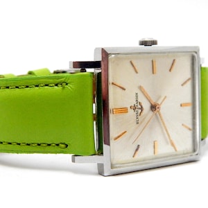 Pu&ograve; includere: Un orologio da polso vintage con quadrante quadrato argentato e lancette e indici dorati. L'orologio presenta un quadrante colorato chiaro e un cinturino in pelle verde brillante. Il quadrante dell'orologio recita "Ulysse Nardin".