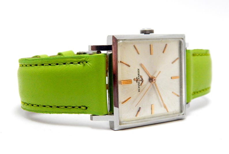 Pu&ograve; includere: Un orologio da polso vintage con quadrante quadrato argentato e lancette e indici dorati. L'orologio presenta un quadrante colorato chiaro e un cinturino in pelle verde brillante. Il quadrante dell'orologio recita "Ulysse Nardin".