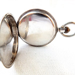K&ouml;nnte beinhalten: Ge&ouml;ffnete silberne Taschenuhr mit ge&ouml;ffnetem Deckel, der das Innenleben zeigt. Die Uhr hat ein rundes Zifferblatt und eine Schlaufe zum Anbringen einer Kette. Die Uhr befindet sich vor einem wei&szlig;en Hintergrund.