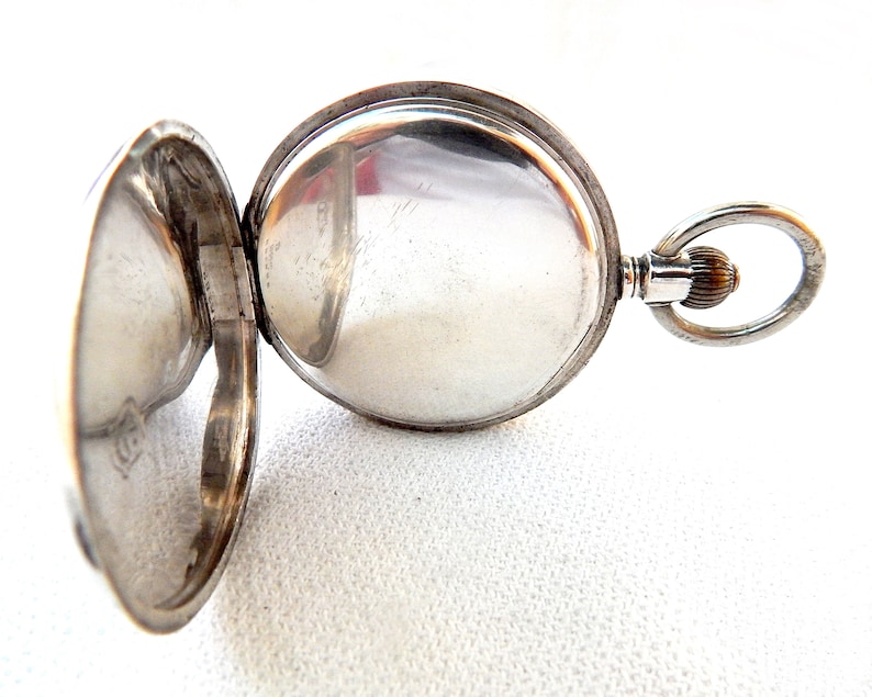 K&ouml;nnte beinhalten: Ge&ouml;ffnete silberne Taschenuhr mit ge&ouml;ffnetem Deckel, der das Innenleben zeigt. Die Uhr hat ein rundes Zifferblatt und eine Schlaufe zum Anbringen einer Kette. Die Uhr befindet sich vor einem wei&szlig;en Hintergrund.