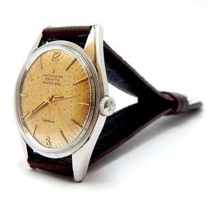 May include: A vintage Zenith Captain automatic wristwatch with a silver case and a brown leather strap. The watch face is a speckled gold colour with gold hands and markers. The watch has the text "CHRONOMETRE ZENITH automatic" and "Captain" on the face.