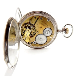 May include: Open silver pocket watch with the back cover ajar, revealing the intricate gold-toned gears and mechanisms inside. The watch face displays the text "CYMA". The watch has a silver loop and a textured crown.