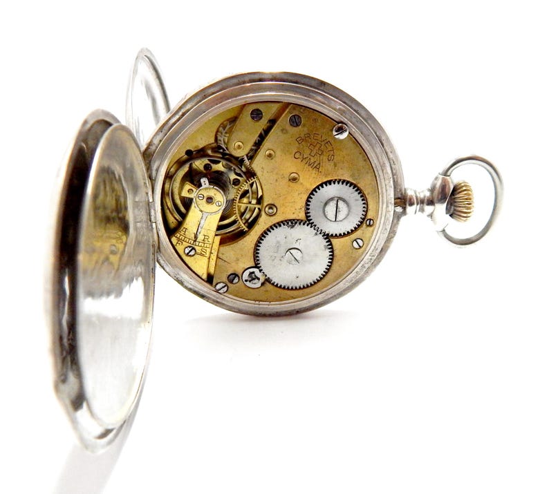 May include: Open silver pocket watch with the back cover ajar, revealing the intricate gold-toned gears and mechanisms inside. The watch face displays the text "CYMA". The watch has a silver loop and a textured crown.