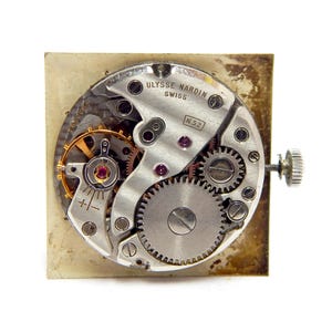 Pu&ograve; includere: Primo piano del meccanismo di un orologio con ingranaggi, gioielli e la scritta "Ulysse Nardin Swiss". Il meccanismo dell'orologio &egrave; posto su uno sfondo quadrato, invecchiato e bianco sporco. I dettagli intricati dei componenti interni sono ben visibili.