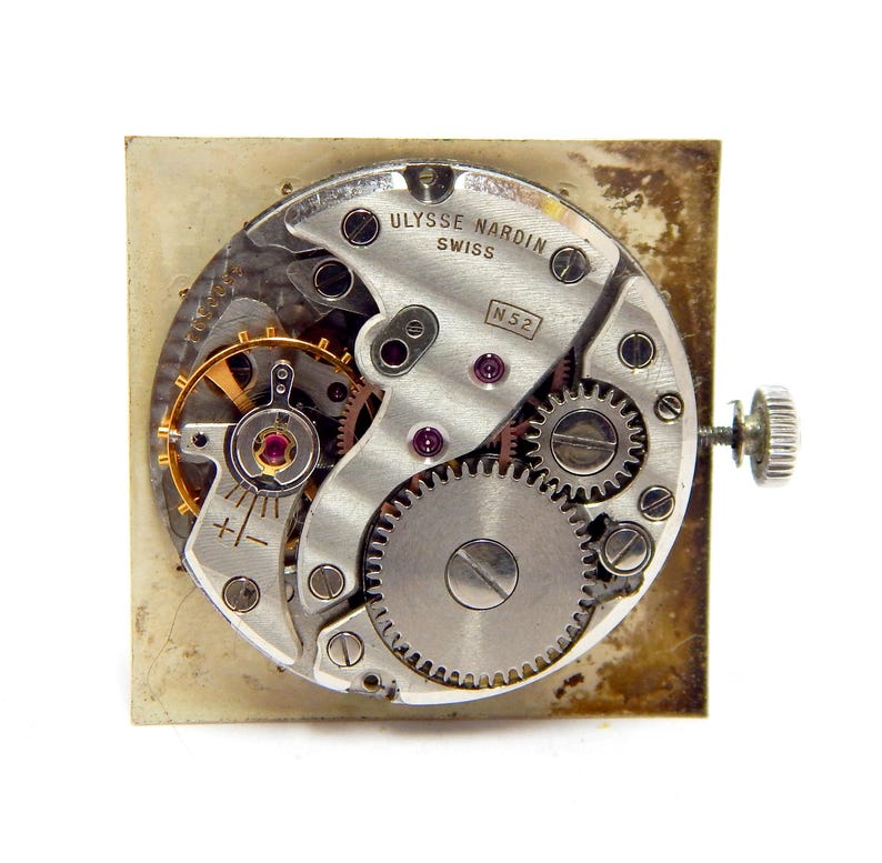 Pu&ograve; includere: Primo piano del meccanismo di un orologio con ingranaggi, gioielli e la scritta "Ulysse Nardin Swiss". Il meccanismo dell'orologio &egrave; posto su uno sfondo quadrato, invecchiato e bianco sporco. I dettagli intricati dei componenti interni sono ben visibili.