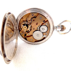 K&ouml;nnte beinhalten: Ge&ouml;ffnete silberne Taschenuhr, die komplizierte mechanische Komponenten zeigt. Das Zifferblatt der Uhr ist freigelegt und zeigt Zahnr&auml;der, Federn und andere interne Mechanismen. Die Uhr ist ge&ouml;ffnet und zeigt das Innenleben.