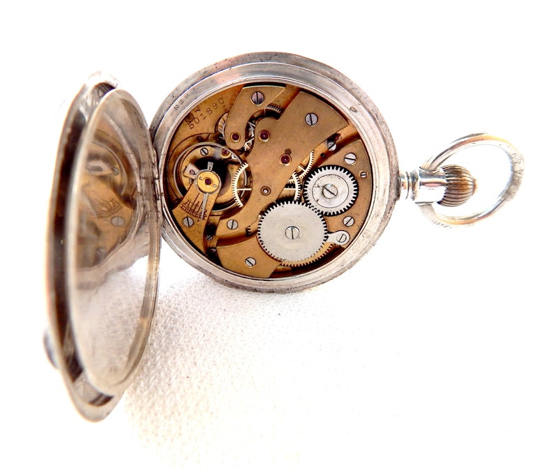 K&ouml;nnte beinhalten: Ge&ouml;ffnete silberne Taschenuhr, die komplizierte mechanische Komponenten zeigt. Das Zifferblatt der Uhr ist freigelegt und zeigt Zahnr&auml;der, Federn und andere interne Mechanismen. Die Uhr ist ge&ouml;ffnet und zeigt das Innenleben.