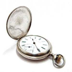 May include: A silver pocket watch with the cover open, revealing a white face with black Roman numerals and a smaller seconds dial. The watch has a gold-colored ring and a silver chain attachment.