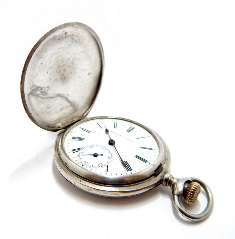 May include: A silver pocket watch with the cover open, revealing a white face with black Roman numerals and a smaller seconds dial. The watch has a gold-colored ring and a silver chain attachment.