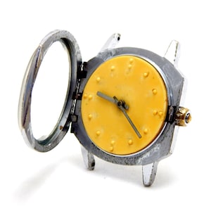 May include: A vintage wristwatch with a silver-colored metal case and a yellow face. The watch face has small, raised dots marking the hours. The watch's protective cover is open, revealing the watch face and hands.