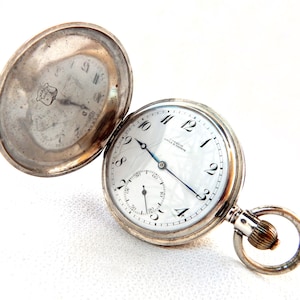 K&ouml;nnte beinhalten: Ge&ouml;ffnete silberne Taschenuhr mit wei&szlig;em Zifferblatt, schwarzen Ziffern und einem kleinen Sekundenzeiger. Die Uhr hat einen silbernen Kettenring und die R&uuml;ckseite ist offen. Auf dem Zifferblatt steht "CHARDIN, COLA & GENEVE".