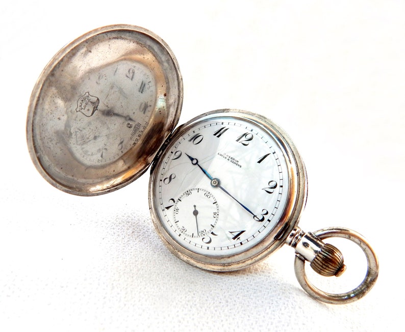 K&ouml;nnte beinhalten: Ge&ouml;ffnete silberne Taschenuhr mit wei&szlig;em Zifferblatt, schwarzen Ziffern und einem kleinen Sekundenzeiger. Die Uhr hat einen silbernen Kettenring und die R&uuml;ckseite ist offen. Auf dem Zifferblatt steht "CHARDIN, COLA & GENEVE".