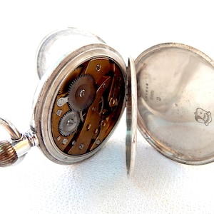 K&ouml;nnte beinhalten: Ge&ouml;ffnete silberne Taschenuhr mit sichtbarem Uhrwerk. Die Uhr hat einen Klappdeckel und eine dekorative Schlaufe. Das Innenleben zeigt messingfarbene Zahnr&auml;der und Schrauben.