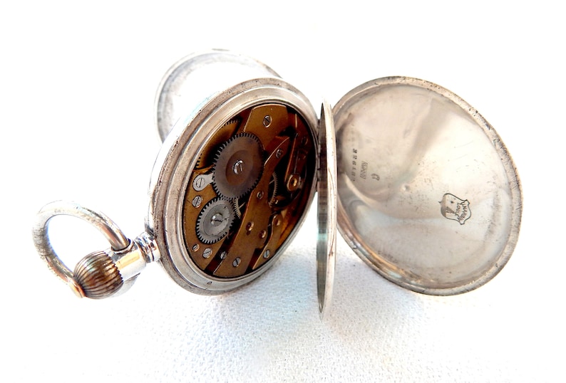 K&ouml;nnte beinhalten: Ge&ouml;ffnete silberne Taschenuhr mit sichtbarem Uhrwerk. Die Uhr hat einen Klappdeckel und eine dekorative Schlaufe. Das Innenleben zeigt messingfarbene Zahnr&auml;der und Schrauben.