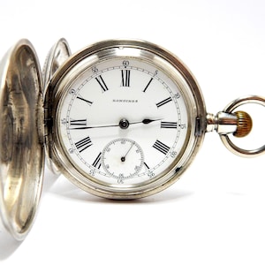 May include: A silver-toned Longines pocket watch with an open hinged cover, a white dial featuring black Roman numerals, a small sub-dial for seconds, and a metallic winding crown with a circular ring attachment at the top of the watch case.