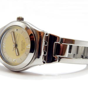 Puede incluir: Un reloj de pulsera Swatch Swiss con una correa de metal plateado y una esfera de color amarillo claro. El reloj tiene una esfera redonda con manecillas e indicadores de hora plateados. El reloj se muestra sobre una superficie blanca.