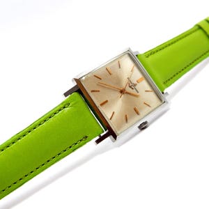 Pu&ograve; includere: Un orologio da polso vintage con quadrante quadrato color oro e cassa color argento. L'orologio presenta un cinturino in pelle verde brillante. Il quadrante dell'orologio ha lancette e indici color oro. Il cinturino &egrave; largo circa 2 cm.