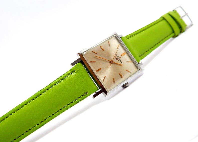 Pu&ograve; includere: Un orologio da polso vintage con quadrante quadrato color oro e cassa color argento. L'orologio presenta un cinturino in pelle verde brillante. Il quadrante dell'orologio ha lancette e indici color oro. Il cinturino &egrave; largo circa 2 cm.