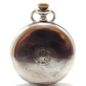 May include: A silver pocket watch with a gold-colored crown and a textured, engraved surface. The watch features a circular monogram on the back. The watch is a classic timepiece.