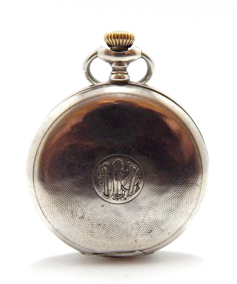May include: A silver pocket watch with a gold-colored crown and a textured, engraved surface. The watch features a circular monogram on the back. The watch is a classic timepiece.