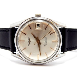 Puede incluir: Un reloj autom&aacute;tico Longines Conquest vintage con esfera plateada, agujas e indicadores dorados, y ventana de fecha a las 3. El reloj tiene caja de acero inoxidable y correa de cuero negro. Un reloj cl&aacute;sico.