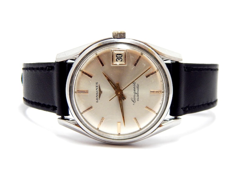 Puede incluir: Un reloj autom&aacute;tico Longines Conquest vintage con esfera plateada, agujas e indicadores dorados, y ventana de fecha a las 3. El reloj tiene caja de acero inoxidable y correa de cuero negro. Un reloj cl&aacute;sico.