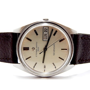 Puede incluir: Un reloj de pulsera vintage Eterna-Matic 3000 con esfera plateada, manecillas negras y correa de cuero marrón. La esfera muestra el día y la fecha, con el texto "SEVENDAY" y "LUN 22".