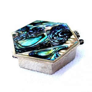 May include: A small, silver-toned hexagonal box with a hinged lid. The lid is inlaid with iridescent abalone shell, displaying shades of blue, green, and purple. The box has a simple, geometric design and a small clasp.