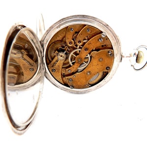 May include: Open silver pocket watch with the cover ajar, revealing the intricate brass gears and mechanisms. The watch has a silver case and a small crown. The watch is set against a white background.