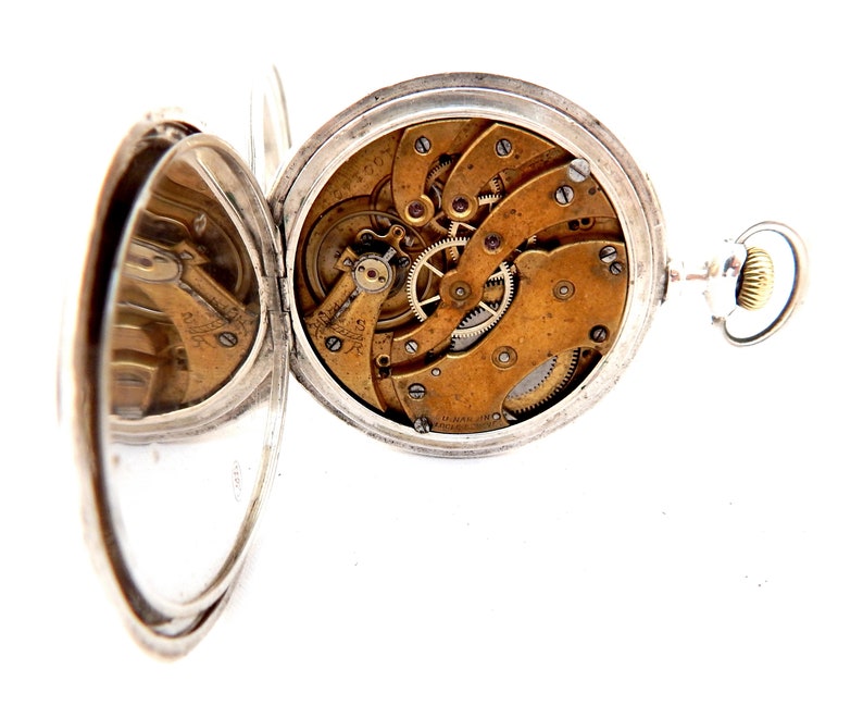 May include: Open silver pocket watch with the cover ajar, revealing the intricate brass gears and mechanisms. The watch has a silver case and a small crown. The watch is set against a white background.
