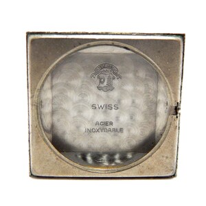 Pu&ograve; includere: Una cassa di orologio vintage color argento con un quadrante circolare trasparente. Il quadrante mostra il testo "SWISS" e "ACIER INOXYDABLE". L'orologio ha una forma rettangolare e un fondello incernierato.