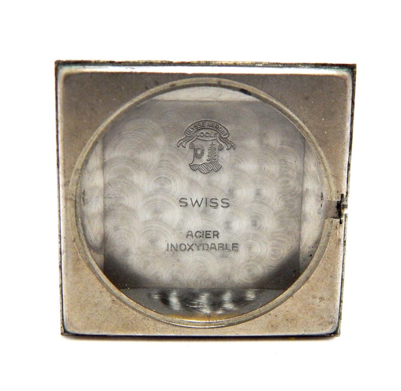 Pu&ograve; includere: Una cassa di orologio vintage color argento con un quadrante circolare trasparente. Il quadrante mostra il testo "SWISS" e "ACIER INOXYDABLE". L'orologio ha una forma rettangolare e un fondello incernierato.