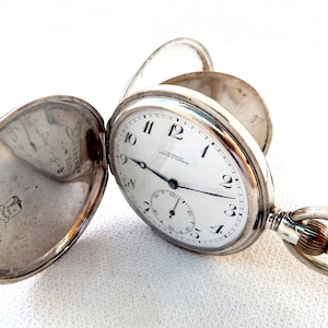 K&ouml;nnte beinhalten: Offene silberne Taschenuhr mit wei&szlig;em Zifferblatt, schwarzen Ziffern und einem Hilfszifferblatt. Die Uhr hat einen Klappdeckel und einen Ring f&uuml;r eine Kette. Auf dem Zifferblatt steht "J. G. Graves, Sheffield & London".
