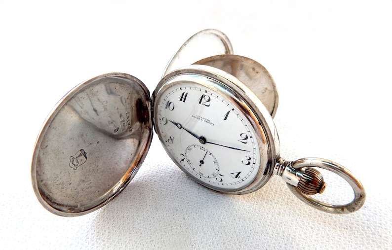 K&ouml;nnte beinhalten: Offene silberne Taschenuhr mit wei&szlig;em Zifferblatt, schwarzen Ziffern und einem Hilfszifferblatt. Die Uhr hat einen Klappdeckel und einen Ring f&uuml;r eine Kette. Auf dem Zifferblatt steht "J. G. Graves, Sheffield & London".