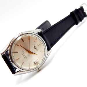 Puede incluir: Un reloj de pulsera autom&aacute;tico Longines Conquest vintage con caja plateada y correa de cuero negro. La esfera del reloj es de color crema claro con agujas e indicadores dorados. El reloj tiene una visualizaci&oacute;n de fecha a las 3 en punto.