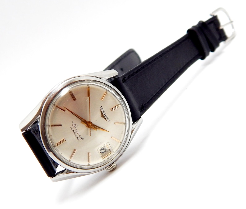 Puede incluir: Un reloj de pulsera autom&aacute;tico Longines Conquest vintage con caja plateada y correa de cuero negro. La esfera del reloj es de color crema claro con agujas e indicadores dorados. El reloj tiene una visualizaci&oacute;n de fecha a las 3 en punto.