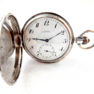 K&ouml;nnte beinhalten: Offene silberne Taschenuhr mit wei&szlig;em Zifferblatt, schwarzen Ziffern und blauen Zeigern. Die Uhr hat ein Hilfszifferblatt und der Markenname "H. Hardin Locle & Geneve" ist auf dem Zifferblatt aufgedruckt. Die Uhr ist ge&ouml;ffnet und zeigt das Innenleben.