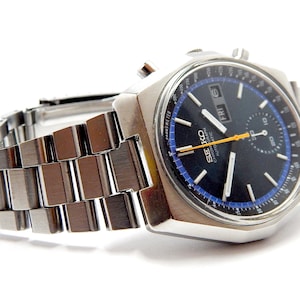 Pode incluir: Um relógio de pulso prateado com mostrador azul escuro e pulseira prateada. O mostrador do relógio tem detalhes brancos e amarelos, com o dia da semana exibido. O relógio é um Seiko Chronograph.
