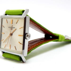 Pu&ograve; includere: Un orologio da polso Ulysse Nardin vintage con quadrante rettangolare argentato, lancette e indici dorati e cinturino in pelle verde. L'orologio ha una cassa argentata e un retro in pelle marrone sul cinturino.