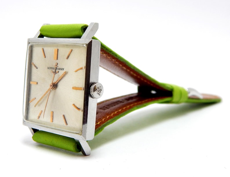 Pu&ograve; includere: Un orologio da polso Ulysse Nardin vintage con quadrante rettangolare argentato, lancette e indici dorati e cinturino in pelle verde. L'orologio ha una cassa argentata e un retro in pelle marrone sul cinturino.