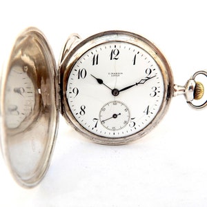 May include: Open silver pocket watch with a white face, black hands, and black Arabic numerals. The watch has a subdial and the brand name "U. NARDIN LOCLE" is visible. The watch is set against a white background.