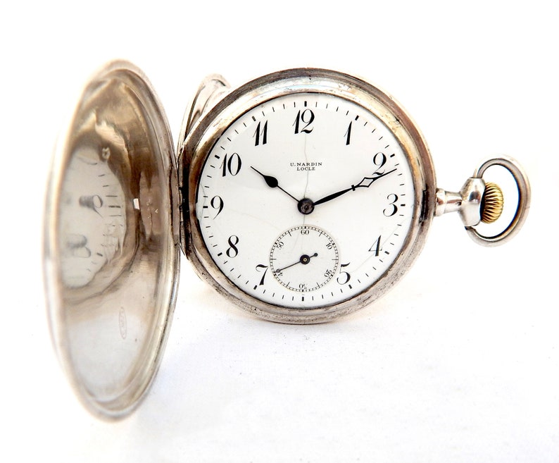 May include: Open silver pocket watch with a white face, black hands, and black Arabic numerals. The watch has a subdial and the brand name "U. NARDIN LOCLE" is visible. The watch is set against a white background.