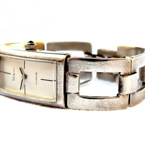 May include: A silver wristwatch with a rectangular face and a silver metal band. The watch face has a white background with black hands and black markings. The watch face also has the text "Valgine" and "Incabloc" printed on it.
