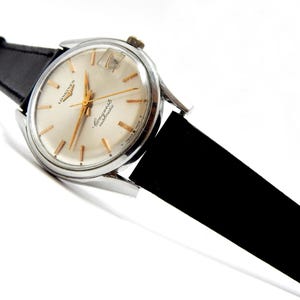 Puede incluir: Un reloj de pulsera autom&aacute;tico Longines Conquest vintage con caja plateada y correa de cuero negra. La esfera del reloj es color crema con agujas e indicadores dorados. El reloj est&aacute; inclinado sobre un fondo blanco.