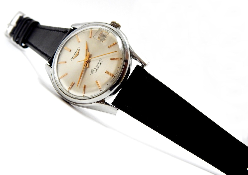 Puede incluir: Un reloj de pulsera autom&aacute;tico Longines Conquest vintage con caja plateada y correa de cuero negra. La esfera del reloj es color crema con agujas e indicadores dorados. El reloj est&aacute; inclinado sobre un fondo blanco.