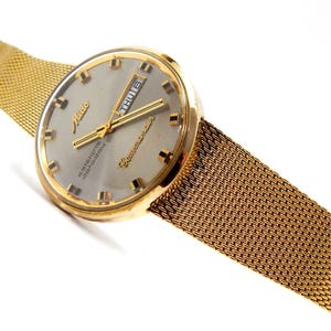 May include: A gold-toned Mido Commander wristwatch with a mesh strap. The watch face is grey with gold hands and markers, showing the day and date. The strap is a woven gold mesh.