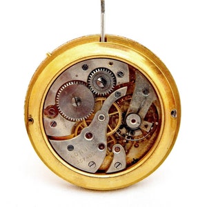 Peut inclure: Un mouvement de montre de couleur dor&eacute;e avec des engrenages complexes et l'inscription "Longines 3574031" visible. Le mouvement de la montre est expos&eacute; et plac&eacute; sur un fond blanc.
