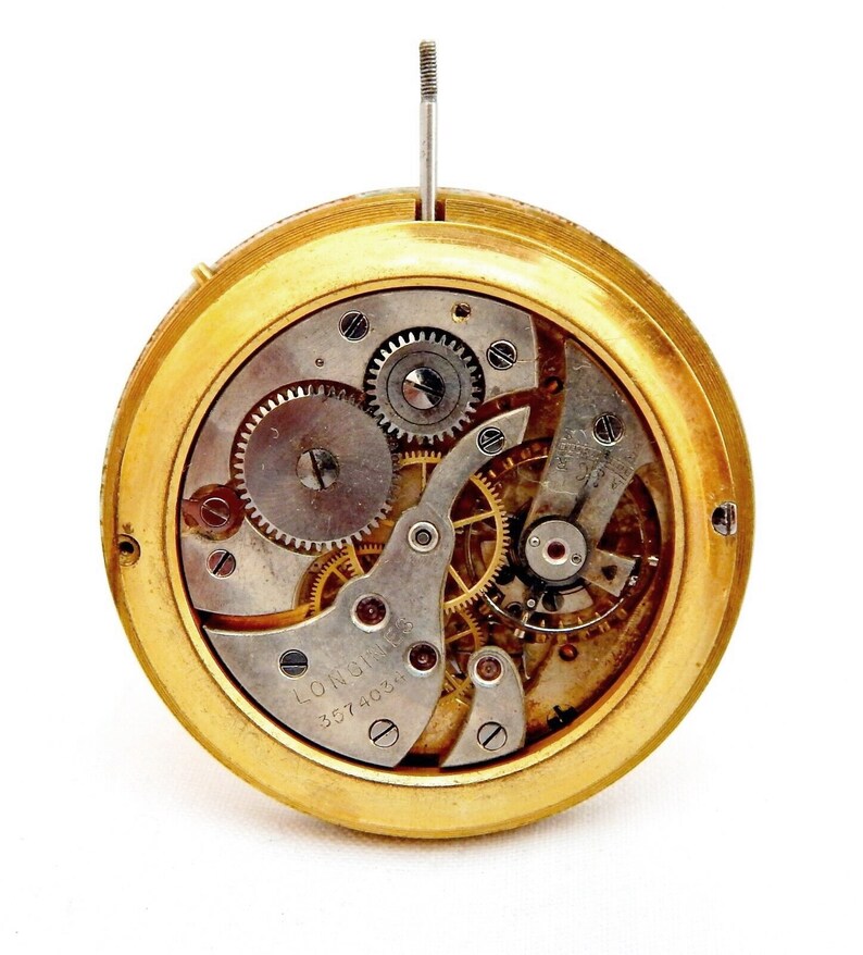 Peut inclure: Un mouvement de montre de couleur dor&eacute;e avec des engrenages complexes et l'inscription "Longines 3574031" visible. Le mouvement de la montre est expos&eacute; et plac&eacute; sur un fond blanc.