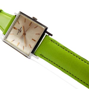 Pu&ograve; includere: Un orologio da polso quadrato con cassa argentata e quadrante chiaro. L'orologio ha lancette e indici color oro. &Egrave; dotato di un cinturino in pelle verde brillante. L'orologio &egrave; angolato su uno sfondo bianco.